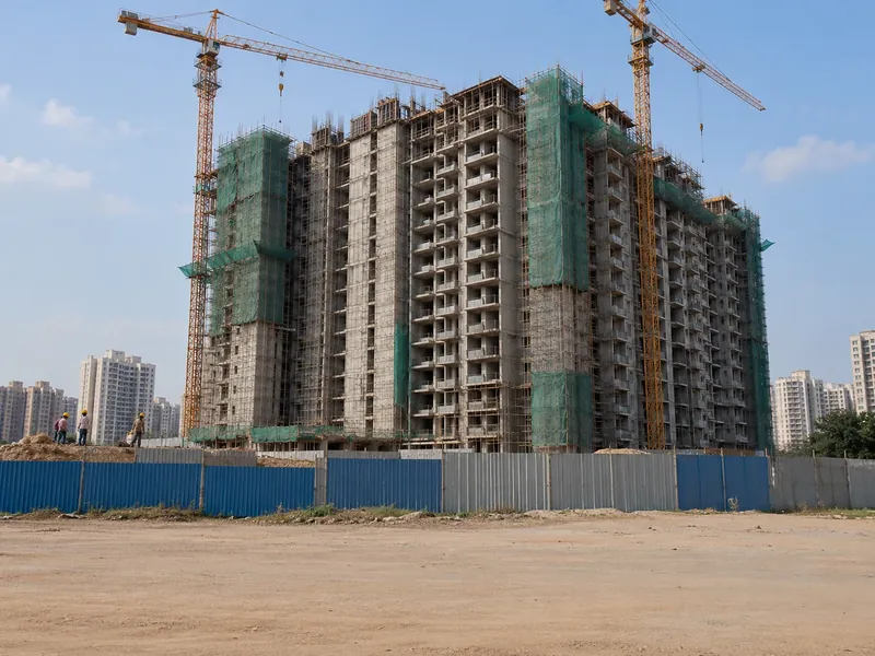 A high-rise residential tower under construction with two tower cranes, green safety netting and a metal-sheet site boundary