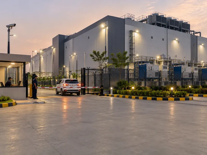 The exterior of a large data centre campus at dusk with a guard cabin, boom barrier, perimeter fence and cooling units